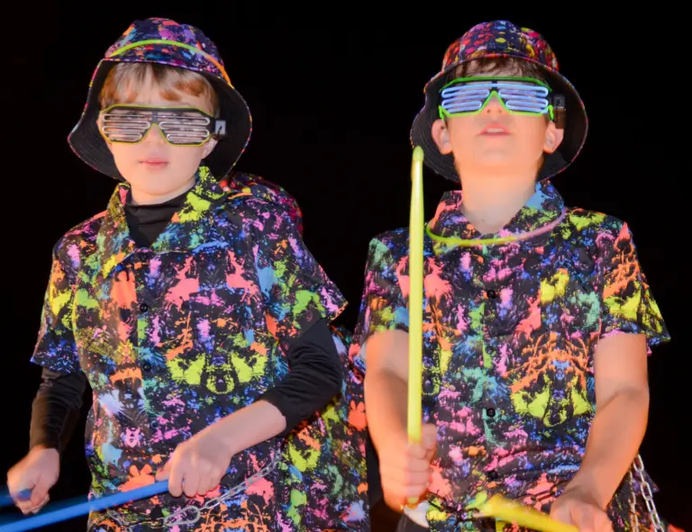 Two boys in neon splatter shirts, bucket hats and slatted party glasses drumming with glow sticks on a carnival float at night