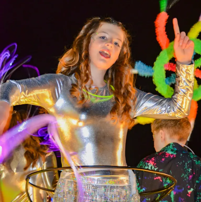 Girl in a shiny silver catsuit and glow necklace dancing and pointing skywards on an illuminated float