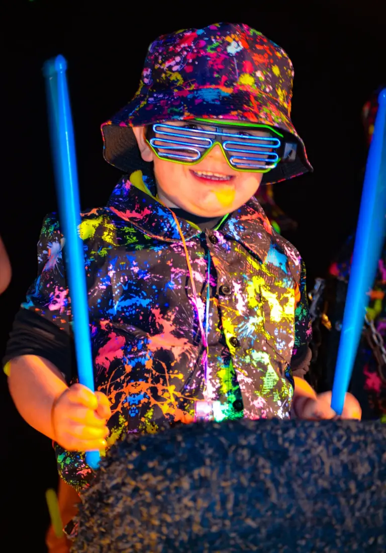 Boy in a neon splatter shirt and slatted glasses drumming with blue glow sticks