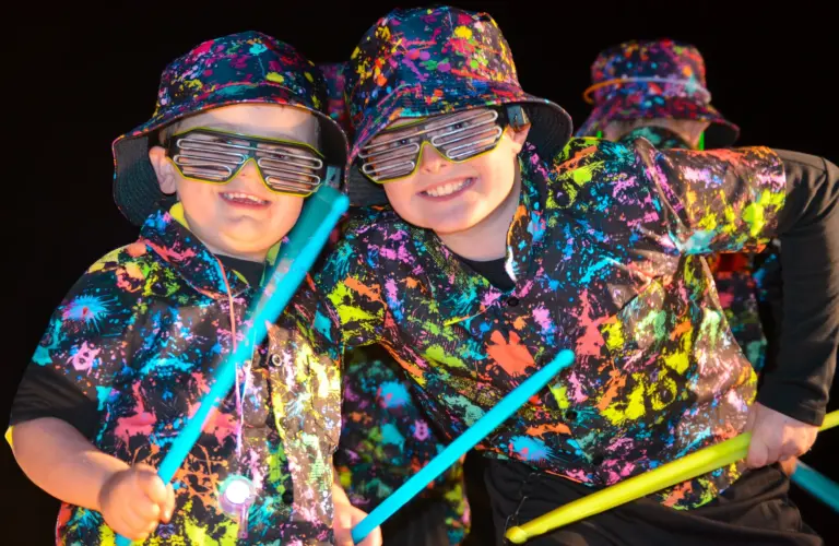 Two grinning boys in neon splatter shirts and party glasses drumming with glow sticks