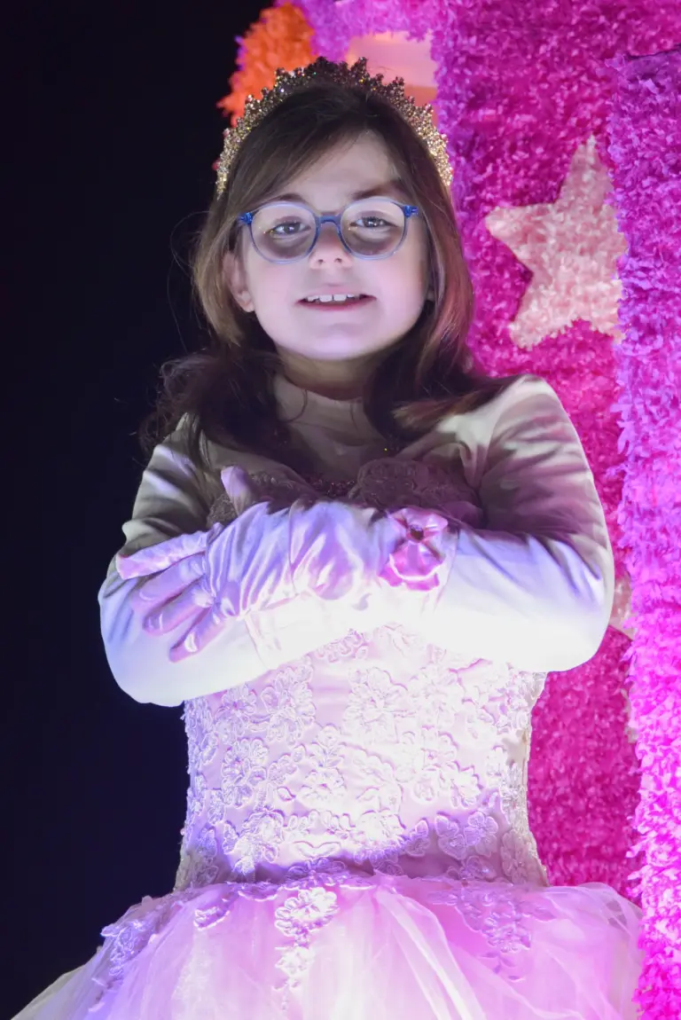 Young girl in glasses and a white lace gown with a tiara on a pink sparkly float