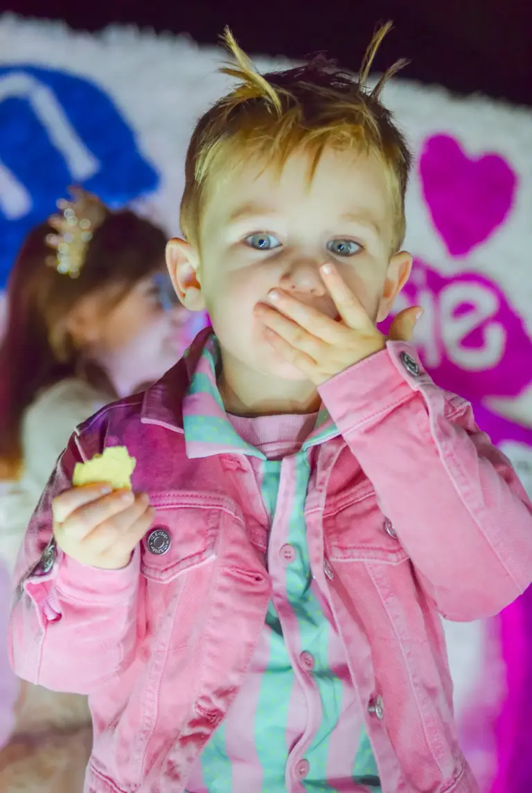 Small boy in a pink denim jacket covering his mouth in surprise on a Barbie themed float