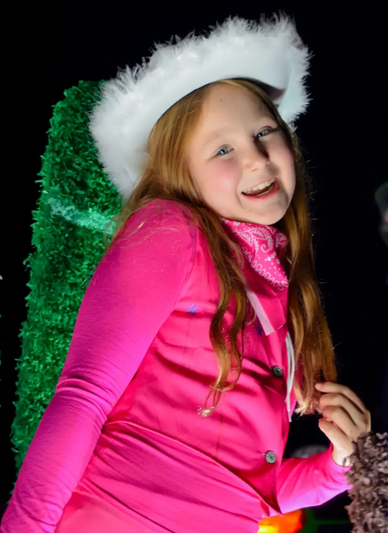 Smiling girl in a pink outfit and white fluffy cowboy hat on an illuminated float
