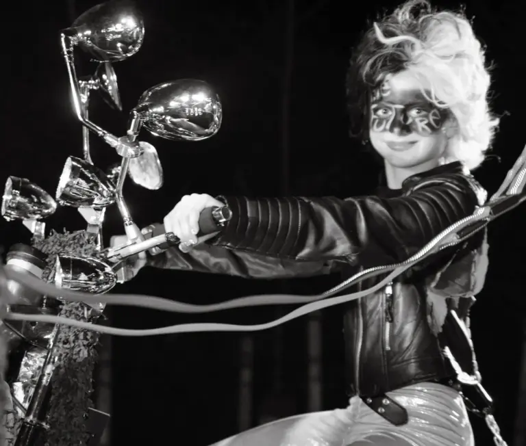 Black and white photo of a costumed rider in a leather jacket and face paint on a chrome motorbike prop