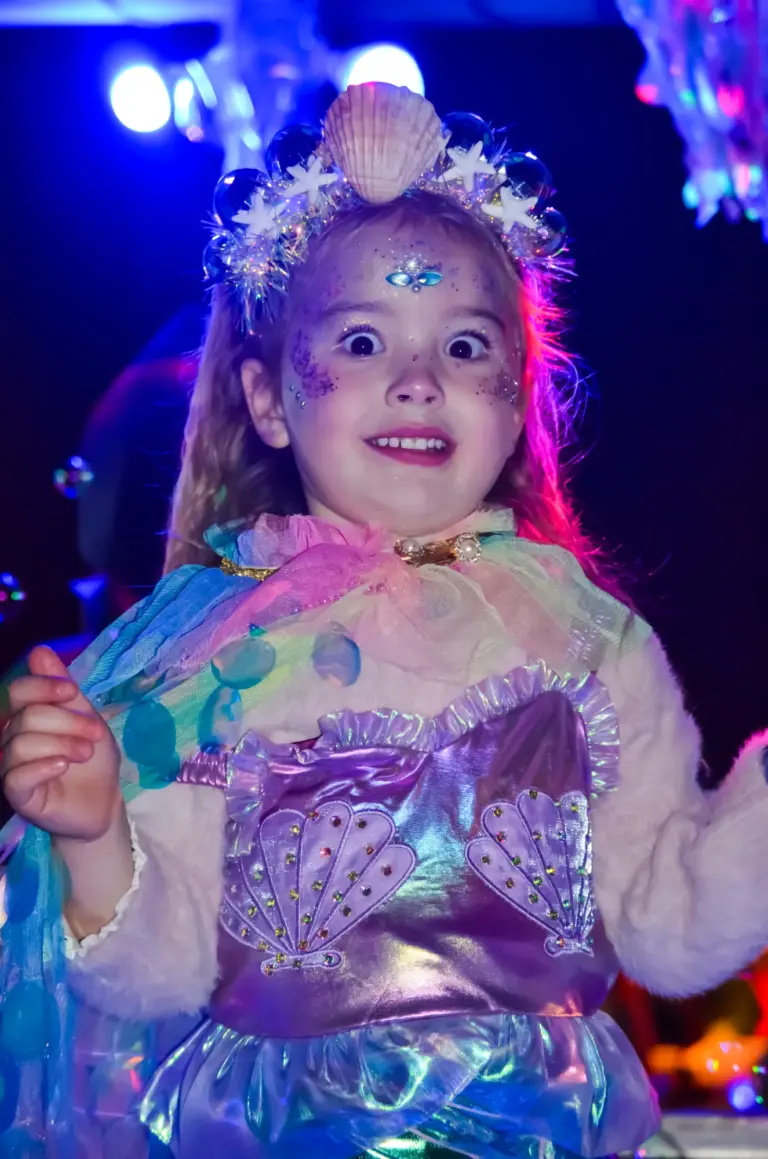 Wide-eyed young girl dressed as a mermaid with a shell crown on a lit-up float