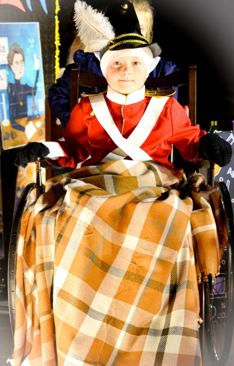 Child dressed as an injured redcoat soldier sitting in a wheelchair with a tartan blanket on a history themed float