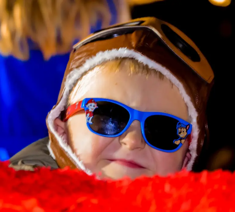 Child in a leather aviator helmet and blue sunglasses peeking over a red feathery float edge