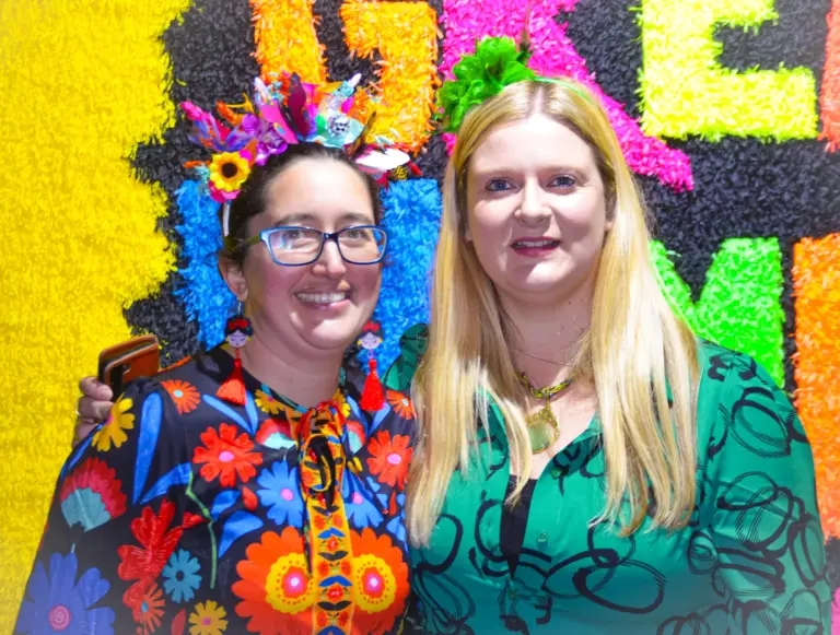 Two women posing in front of a crepe-paper float, one dressed as Frida Kahlo with a flower crown