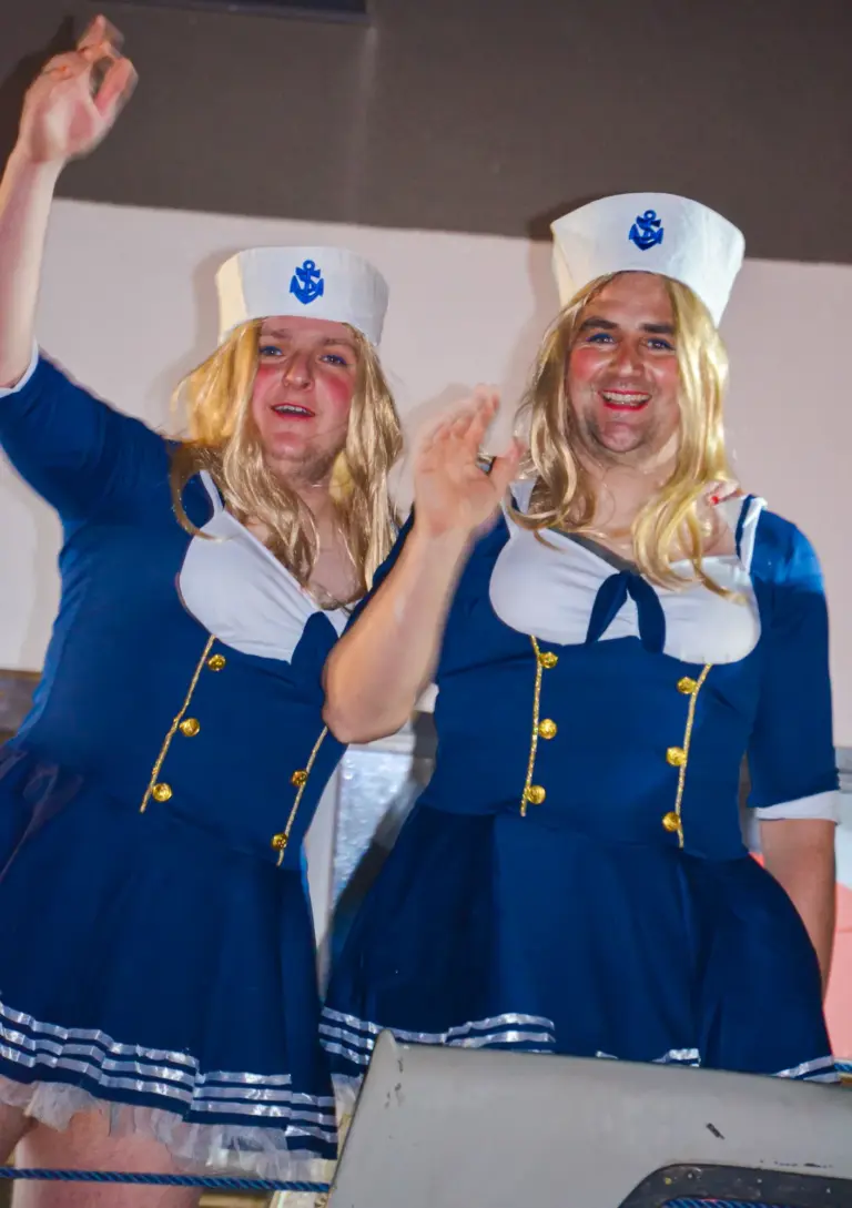 Two men dressed as sailor girls in blonde wigs and blue dresses waving from a boat float