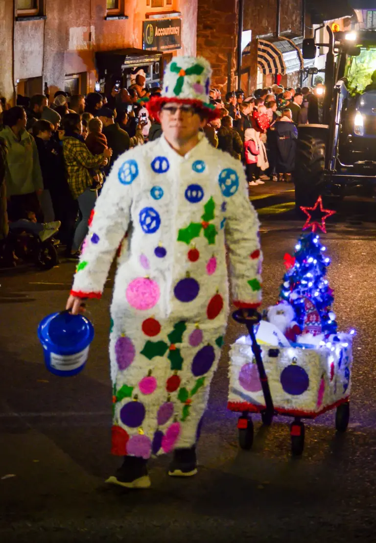 Walking entrant in a white polka-dot costume pulling a cart with a lit Christmas tree