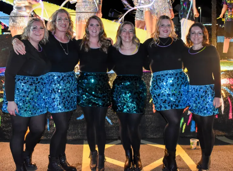 Six women in black tops and blue sequin skirts posing together in front of a float