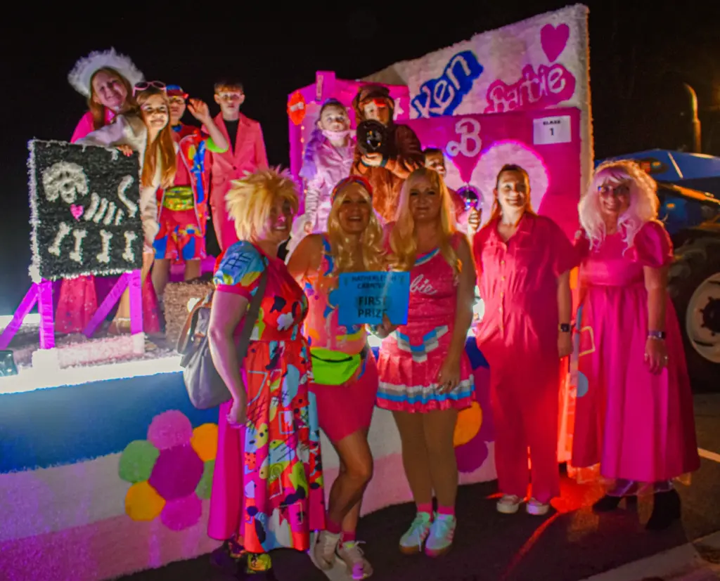 Group in bright pink outfits and wigs posing in front of the Barbie and Ken float