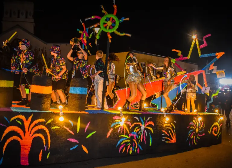 Performers in neon costumes and silver outfits dancing on an illuminated float decorated with fireworks designs