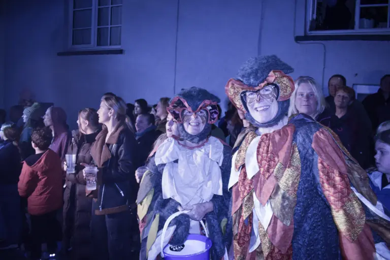 Costumed figures in masks and patchwork robes among the evening crowd