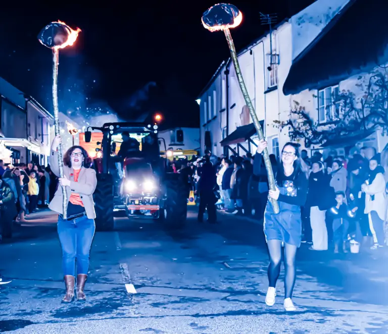 Two women carrying flaming torches through the street ahead of a tractor