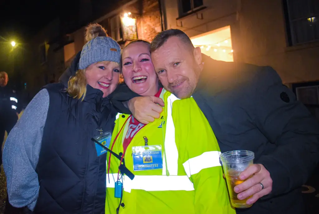 Three people including a marshal in a hi-vis jacket hugging and smiling at night
