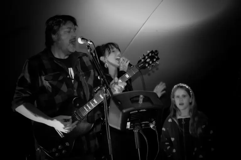 Black and white photo of a band performing with guitar and vocals as a child watches