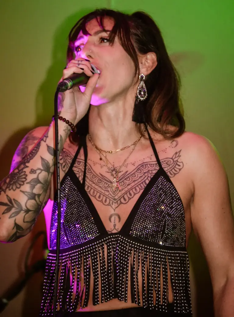 Singer with tattoos and a sparkly fringed top performing under coloured stage lights