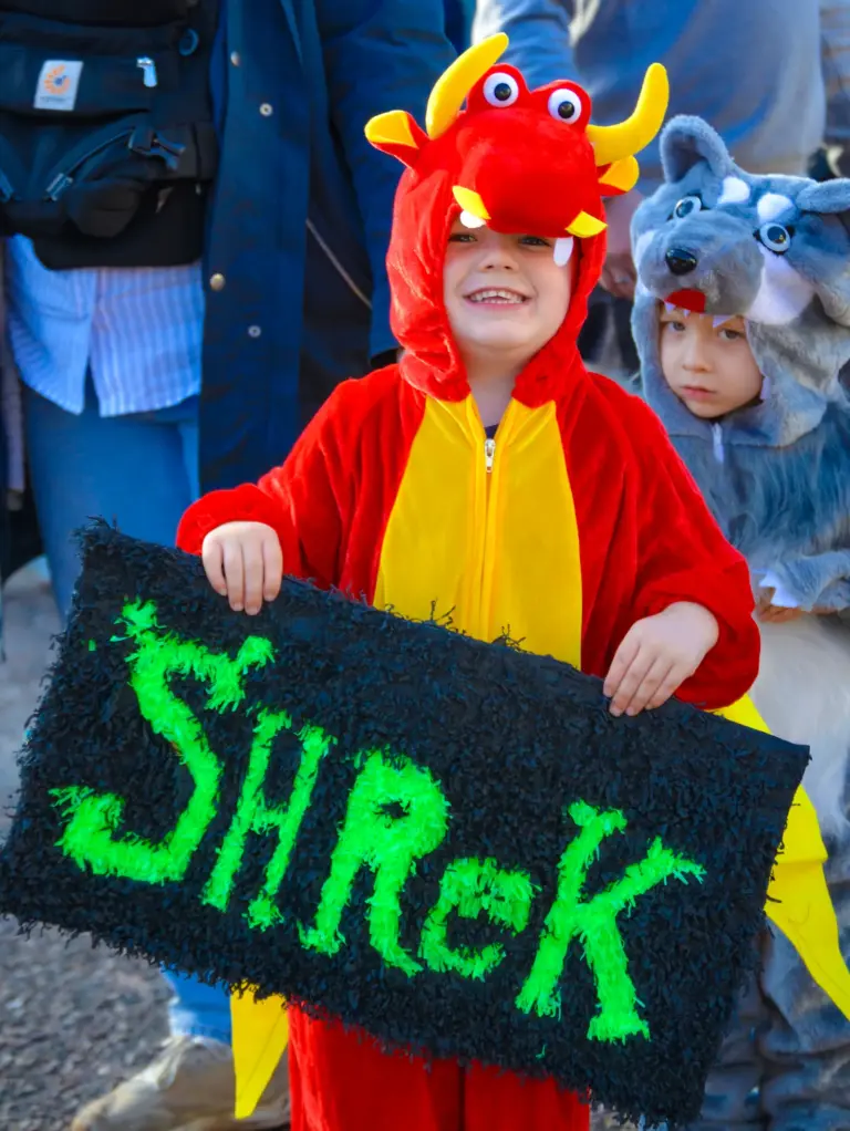 Child in a dragon costume holding a green glittery Shrek sign