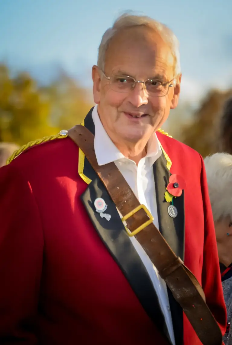 Smiling older gentleman in a red ceremonial jacket with a sash and poppy