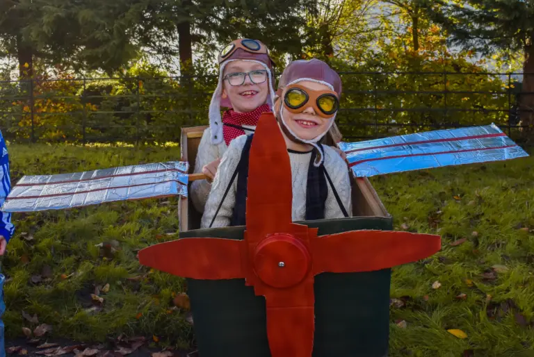 Two children in aviator helmets and goggles in a homemade cardboard aeroplane with a red propeller