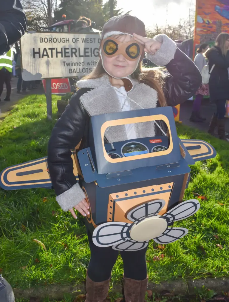 Child in aviator goggles wearing a cardboard aeroplane costume by the Hatherleigh sign