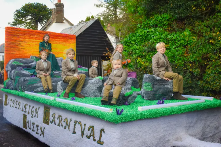 Children in tweed outfits sitting on the Hatherleigh Carnival Queen float decorated with rocks and a sunset backdrop