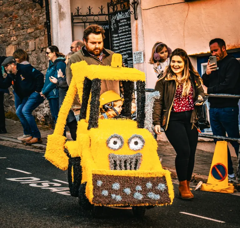 Toddler in a yellow crepe-paper digger pushed along the street by the crowd