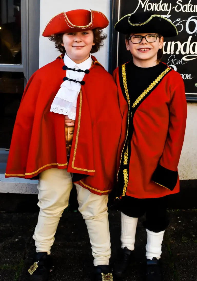 Two smiling young town criers in red coats and tricorn hats standing outside a shop