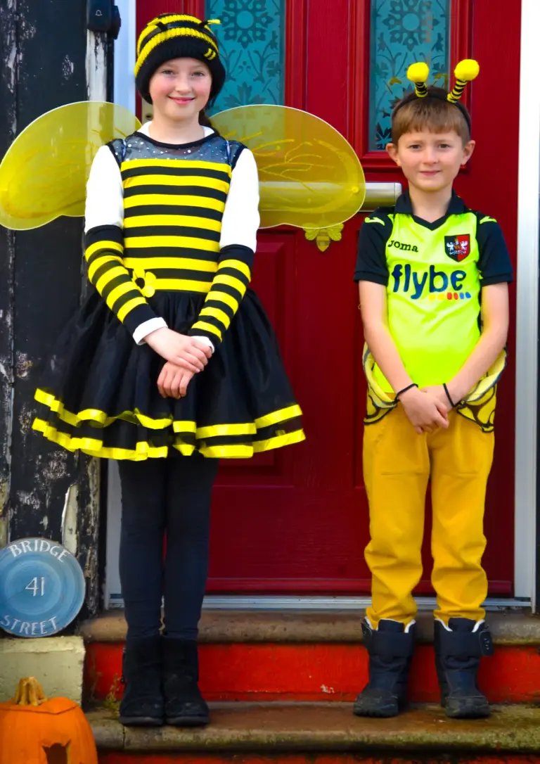Girl in a bee costume and boy in bee antennae on a red front door step