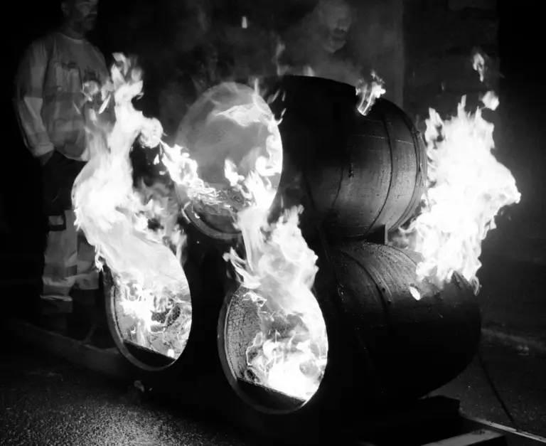 Black and white photo of three tar barrels bursting into flame on a sledge as volunteers stand by
