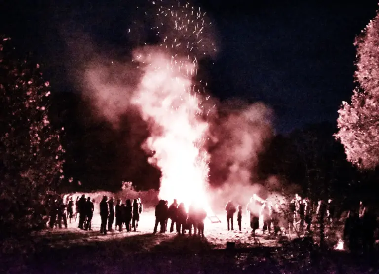 Onlookers silhouetted around the bonfire as white smoke and sparks rise into the night sky