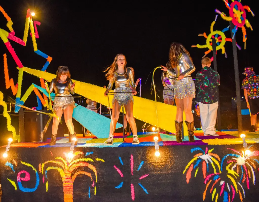 Dancers in silver outfits with hula hoops on a brightly lit neon carnival float at night