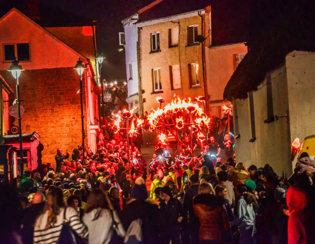 Crowds pack a Hatherleigh street at night as the torchlit procession passes with blazing fire wheels and torches, lighting the buildings red and orange