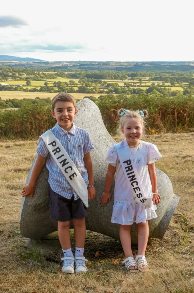 Noah and Mabel Sluggett, Hatherleigh Carnival Prince and Princess 2026, smiling in their Prince and Princess sashes beside a granite sculpture overlooking rolling Devon fields