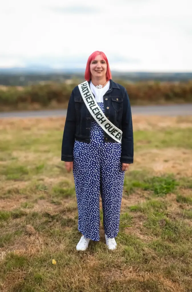 Emma Alford, Hatherleigh Carnival Queen 2026, smiling in a denim jacket and spotted jumpsuit with her Hatherleigh Queen sash, standing in a field with Devon countryside behind