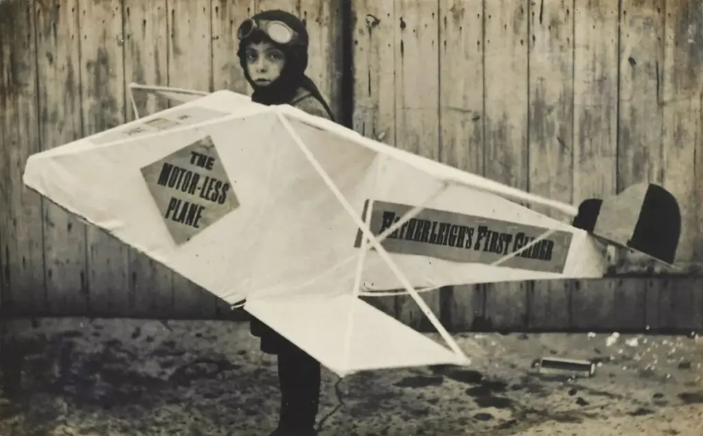 Archive photo of a child in flying goggles wearing a home-made plane costume labelled Hatherleigh's First Glider, The Motor-less Plane