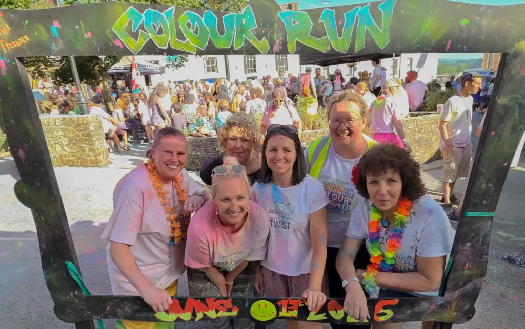 Six smiling members of the Hatherleigh Carnival Committee covered in coloured powder, posing inside a giant painted Colour Run photo frame in the town centre