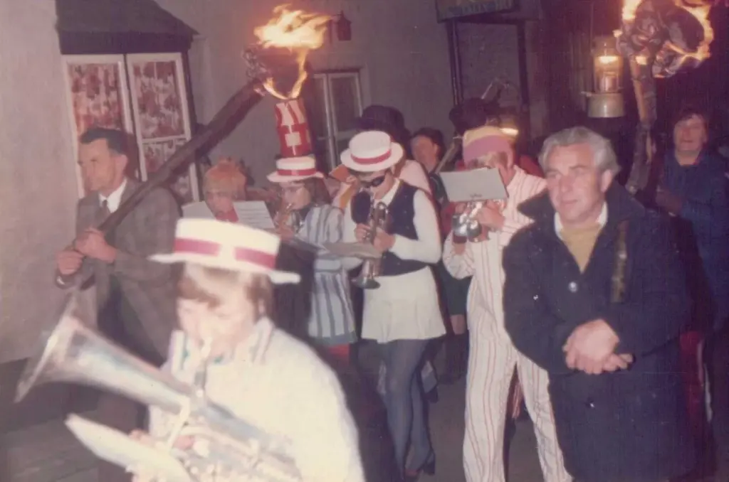 Vintage photo of the Hatherleigh Jazz Band marching through the streets at night in boaters and fancy dress, playing trumpets and a euphonium, with men carrying flaming torches