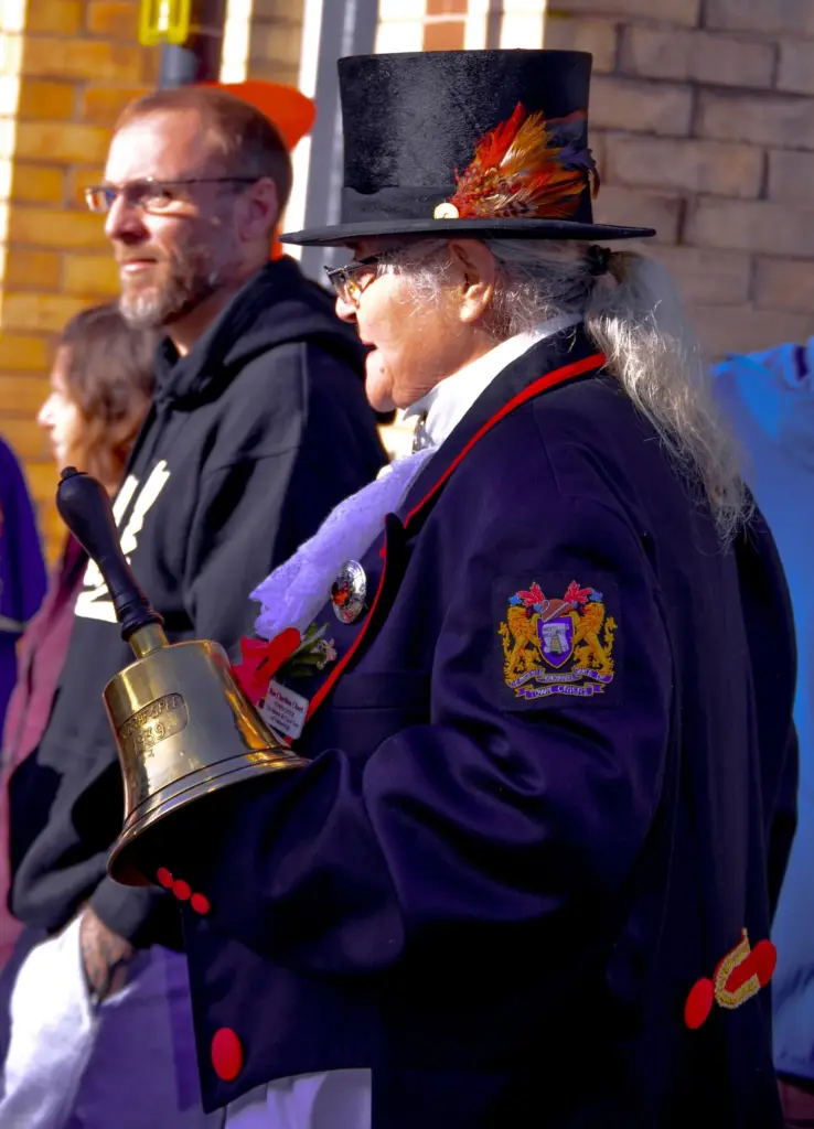 Hatherleigh's town crier in a feathered top hat and red-trimmed coat, holding a brass bell