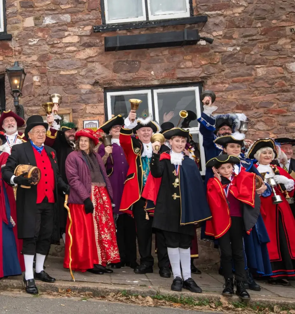 Town criers in colourful ceremonial coats and tricorn hats ringing their bells outside a stone pub in Hatherleigh, including two young criers
