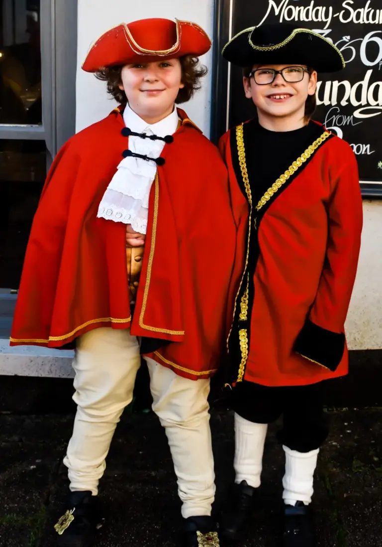 Two smiling junior town criers in tricorn hats, one in a red cape and lace jabot, the other in a red gold-braided coat