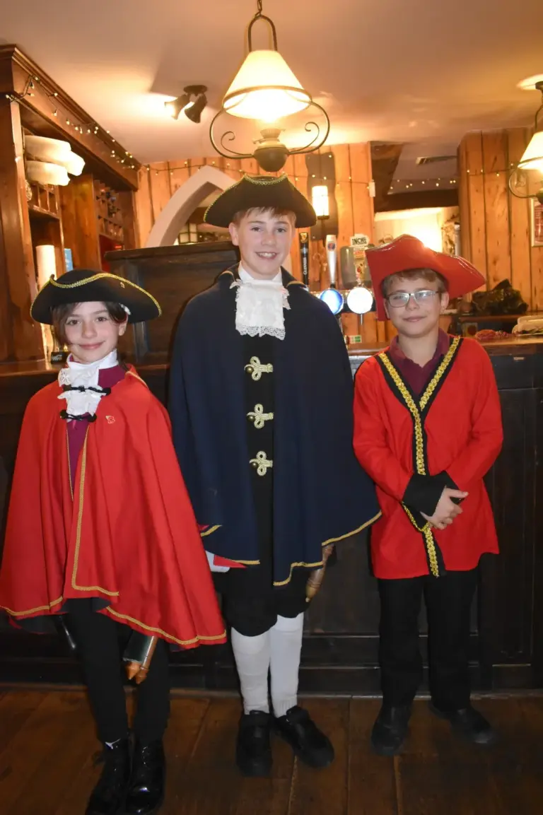 Three junior town criers in tricorn hats, capes and coats smiling together inside the pub