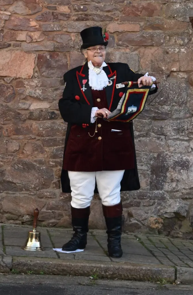 Ros Charlton-Chard, Hatherleigh Town Crier, smiling in a black top hat, red-trimmed tailcoat, lace jabot and white breeches, holding an embroidered scroll with a brass bell at her feet