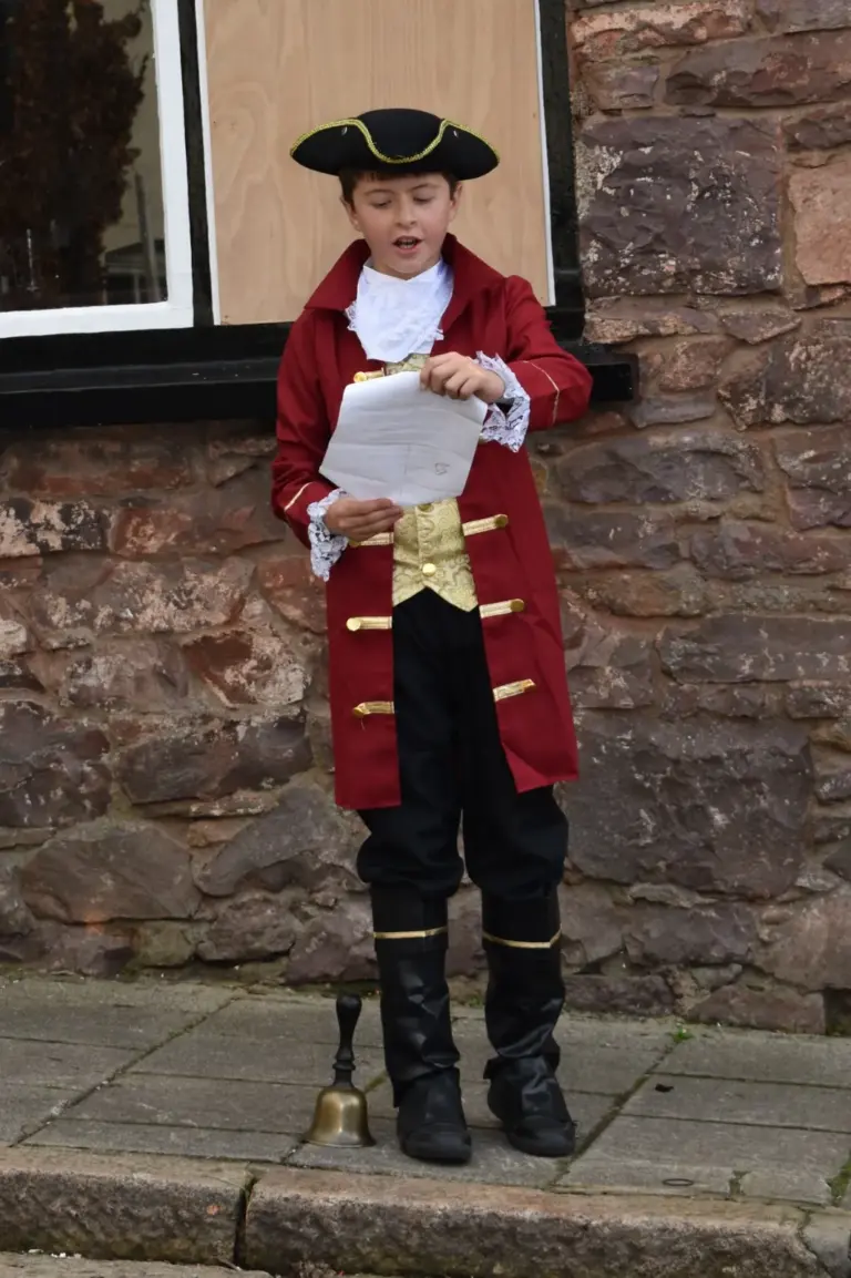Young town crier in a black tricorn hat, burgundy frock coat with gold buttons and a gold waistcoat reading his cry from a sheet of paper