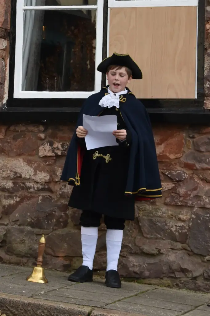 Young town crier in a black tricorn hat, navy cape with gold trim, lace jabot and white stockings reading his cry, with a brass bell beside him