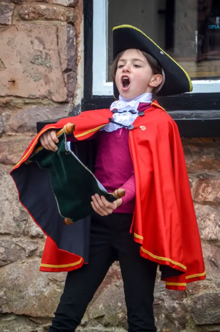 Young town crier in a black tricorn hat, red satin cape and lace jabot reading a cry from a green scroll