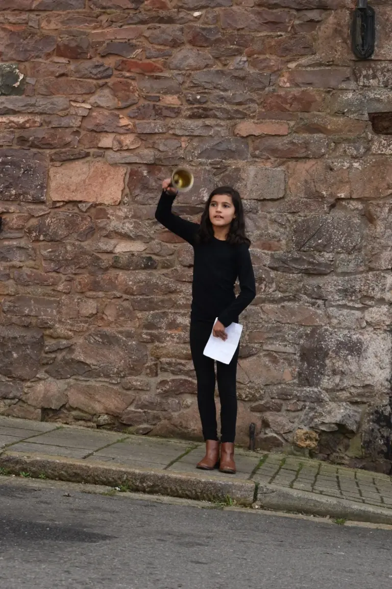 Young town crier dressed in black raising a brass handbell above her head, holding her cry in the other hand, in front of a stone wall